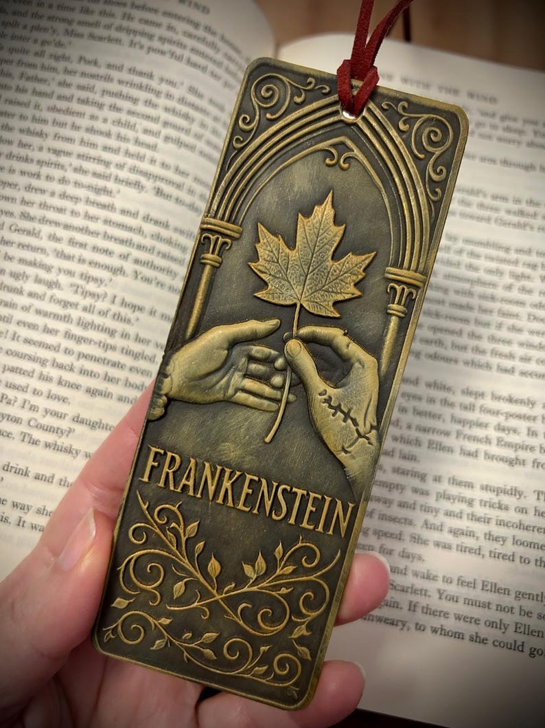 May include: Bronze-colored metal bookmark featuring the word "FRANKENSTEIN" and an illustration of a leaf held by two hands. The bookmark has an arched top and a red cord for easy placement in a book.