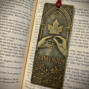 May include: A bronze-colored metal bookmark featuring a maple leaf held by two hands, with the word "FRANKENSTEIN" below. The bookmark has an arched top and a red cord for placement in a book.