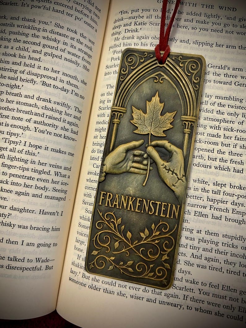 May include: A bronze-colored metal bookmark featuring a maple leaf held by two hands, with the word "FRANKENSTEIN" below. The bookmark has an arched top and a red cord for placement in a book.