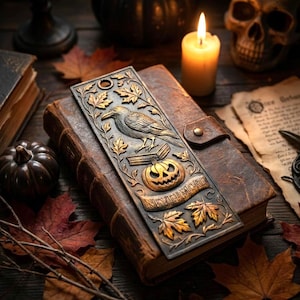 May include: A metal bookmark with a raven, pumpkin, and leaf design, resting on a leather-bound book. The bookmark has the word "NEVERMORE" on it. A lit candle, skull, and autumn leaves are in the background.