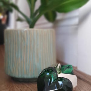 May include: A heart-shaped, dark green perfume bottle with silver detailing rests on a wooden surface. A light green ceramic planter with a plant is in the background. The image has a blurred effect.
