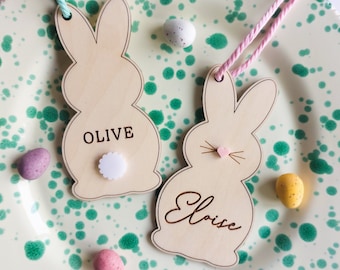 May include: Two wooden bunny-shaped name tags, one reading "Olive" and the other "Eloise", rest on a speckled plate with Easter eggs. A colorful pom-pom garland and decorative eggs surround the plate, creating a festive Easter theme.