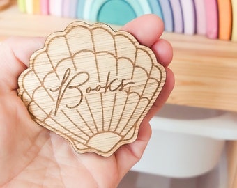 May include: A wooden, seashell-shaped label with the word "Books" engraved on it. The label is held in a hand, with a rainbow-colored wooden arch in the background. The label is designed for organizing books.