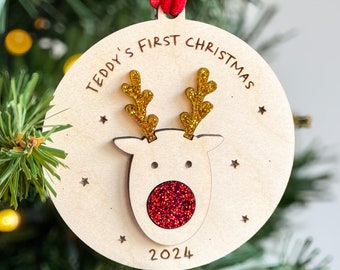 May include: Wooden Christmas ornament with a reindeer design. The ornament has gold glitter antlers and a red glitter nose. Text on the ornament reads "TEDDY'S FIRST CHRISTMAS 2024". A red ribbon is attached for hanging.