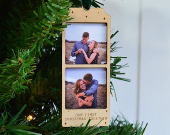 May include: A wooden Christmas ornament with two photo slots, each containing a couple's photo. The ornament has the text "OUR FIRST CHRISTMAS TOGETHER" and is suspended by a green ribbon. The ornament is hanging on a Christmas tree.