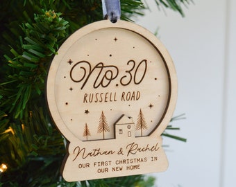 May include: A wooden Christmas ornament shaped like a snow globe, hanging on a green Christmas tree. The ornament has the text "No. 30 Russell Road" and "Nathan & Rachel" with the phrase "Our first Christmas in our new home."