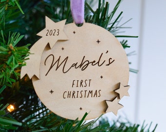 May include: A wooden Christmas ornament with the text "Mabel's First Christmas" and the year "2023". The ornament is round with star accents and a purple ribbon for hanging. It is displayed on a Christmas tree.