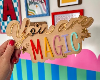 May include: Wooden sign with the words "You are MAGIC" in gold and colorful letters. The sign is held in front of a wall with framed art and a colorful striped and cloud-shaped border.