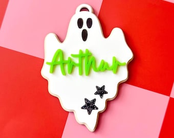 May include: A white ghost-shaped ornament featuring a cutout at the top for hanging. The ghost has black oval eyes and a mouth, with the name Arthur in raised lime green script lettering across the center and two black glittery stars below.