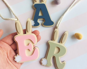 May include: Wooden Easter bunny pendants spelling "EAR", with the letters in pink, blue, and green. Each pendant has a bunny ear design and a white string for hanging. Easter egg candies are scattered around the pendants.