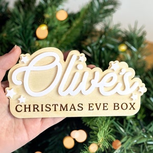 May include: A wooden Christmas Eve box, featuring the name "Olive's" in white script, and the words "CHRISTMAS EVE BOX" in a serif font. The box is light wood-colored with white star accents.