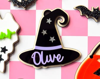 May include: A black acrylic witch hat ornament featuring a light purple band and the name Olive in light purple script. The hat is decorated with three silver glitter stars and includes a small hole at the top for hanging, displayed among other Halloween-themed shapes.