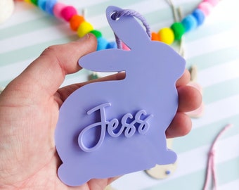 May include: A lavender bunny-shaped tag with the name "Jess" in cursive script. The tag has a hole at the top with a lavender string. A colorful pom-pom garland is in the background. Other Easter-themed tags are also visible.