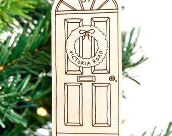 May include: Wooden Christmas ornament shaped like a door, with the number 57 above the door. The door has a wreath that says "Victoria Road" and the text "The Watkinsons" below the door. The ornament hangs from a green ribbon.
