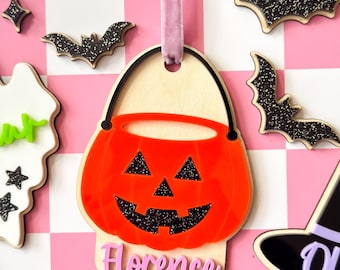 May include: A hanging ornament shaped like a jack-o'-lantern bucket with a bright orange acrylic front, black glitter eyes and mouth, and a light wood backing. The name Florence is written in pink script at the bottom, suspended by a mauve ribbon.