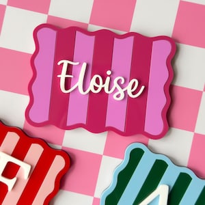 May include: A rectangular plaque with wavy edges featuring vertical stripes in shades of magenta and light pink. The name Eloise is written in white cursive script across the center. Two additional plaques with letters F and A are visible.