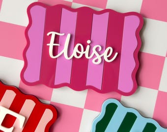 May include: A rectangular plaque with wavy edges featuring vertical stripes in shades of magenta and light pink. The name Eloise is written in white cursive script across the center. Two additional plaques with letters F and A are visible.