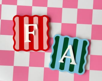May include: Two square coasters with wavy edges. One coaster features red and light pink vertical stripes with a white letter F. The other coaster features dark green and light blue vertical stripes with a white letter A centered on the surface.