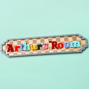 May include: A rectangular wooden sign with a light brown and tan checkered pattern, featuring the text "Arthur's Room" in raised, multi-colored letters. The sign is framed by a silver glitter border with decorative scalloped edges.