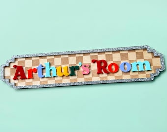 May include: A rectangular wooden sign with a light brown and tan checkered pattern, featuring the text "Arthur's Room" in raised, multi-colored letters. The sign is framed by a silver glitter border with decorative scalloped edges.