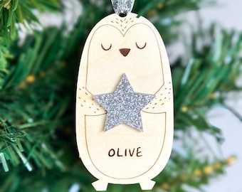 May include: Wooden Christmas ornament shaped like a penguin holding a silver glitter star. The ornament has the name "OLIVE" printed on it and is suspended by a silver ribbon. The ornament is hanging in front of a green Christmas tree.