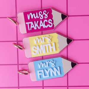 May include: Three pencil-shaped acrylic keychains with glitter edges and gold-tone metal rings. Each features a colored body with white 3D lettering. The top reads miss TAKACS in pink, the middle reads mrs SMITH in yellow, and the bottom reads mrs FLYN in blue.
