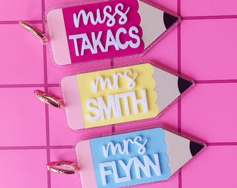 May include: Three pencil-shaped acrylic keychains with glitter edges and gold-tone metal rings. Each features a colored body with white 3D lettering. The top reads miss TAKACS in pink, the middle reads mrs SMITH in yellow, and the bottom reads mrs FLYN in blue.
