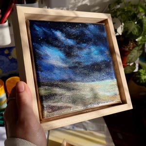 May include: A small framed painting of a night sky with blue and purple clouds. The sky is filled with white stars. The bottom of the painting shows a field of green and brown grass.