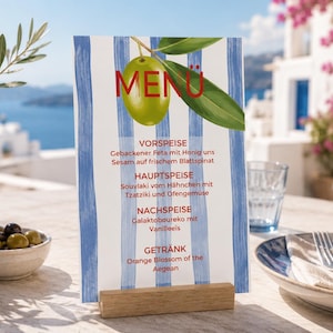 Könnte beinhalten: Eine vertikale Menükarte mit blauen vertikalen Streifen und einer grünen Olivenillustration oben. Der Text lautet MENÜ, VORSPEISE, Gebackener Feta mit Honig uns Sesam auf frischem Blattspinat, HAUPTSPEISE, Souvlaki vom Hähnchen mit Tzatziki und Ofengemüse, NACHSPEISE, Galaktoboureko mit Vanilleeis, GETRÄNK, Orange Blossom of the Aegean.