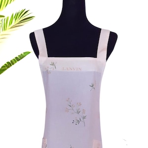 Könnte beinhalten: Eine hellfarbene Schürze mit floralem Design. Die Schürze hat einen quadratischen Ausschnitt und zwei Taschen. Das Wort "LANVIN" ist oben aufgedruckt. Sie ist mit beigefarbenen Blumen und grünen Blättern verziert.