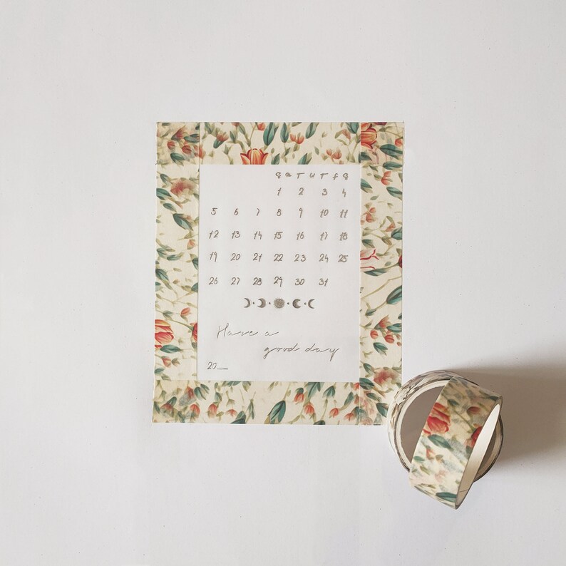 May include: A rectangular calendar sheet featuring a floral border with red tulips and green leaves. The center displays a monthly grid with numbers 1 through 31, moon phase icons, and the handwritten text Have a good day. A roll of matching floral tape sits nearby.