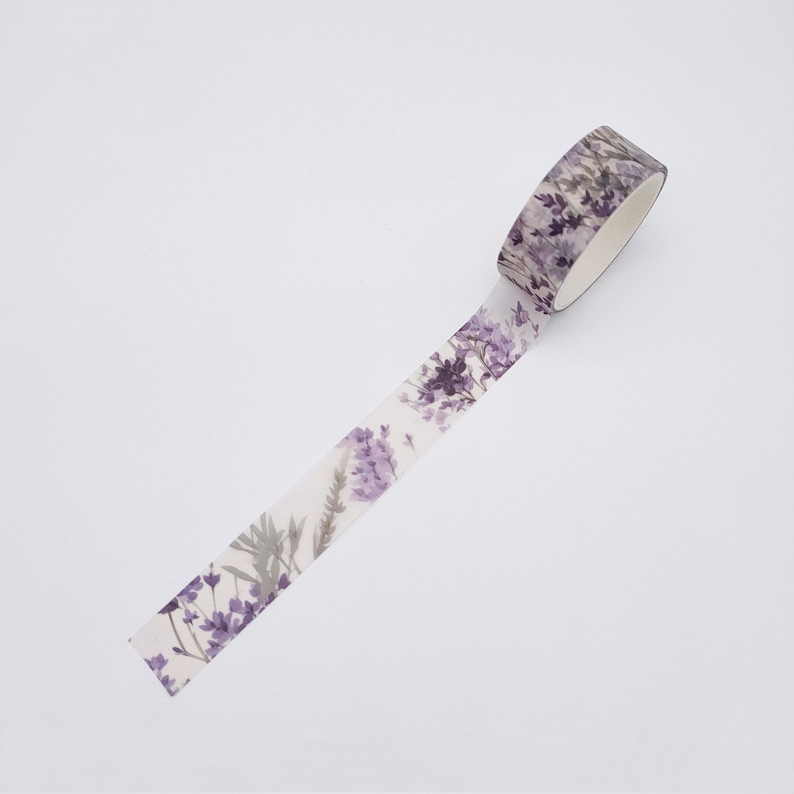 May include: A roll of decorative adhesive tape featuring a repeating watercolour-style pattern of purple lavender sprigs and green stems on a translucent white background. The tape is partially unrolled, displaying the floral botanical design.