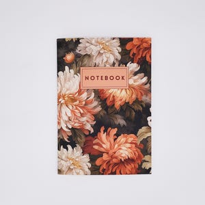 May include: A rectangular notebook featuring a dark background covered in a dense pattern of large, layered chrysanthemum flowers in shades of cream, peach, and burnt orange. A central peach-colored rectangular label displays the word NOTEBOOK.