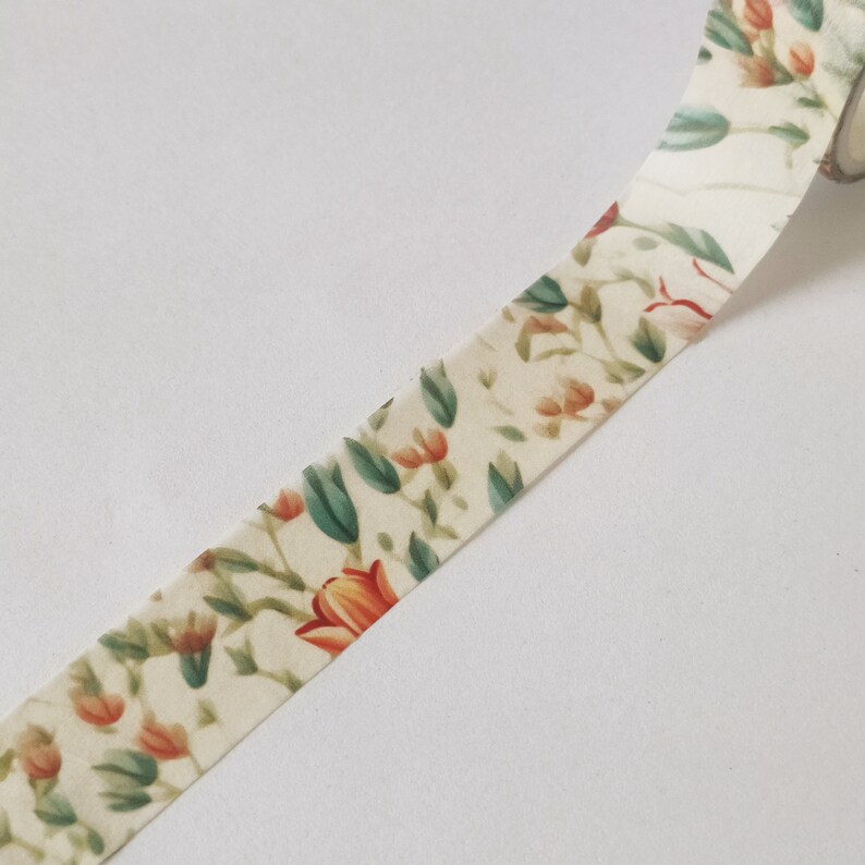 May include: A roll of adhesive tape featuring a cream-colored background with a repeating botanical pattern of orange flowers and muted green leaves. The tape is partially unrolled, displaying the floral print across its length in a continuous design.