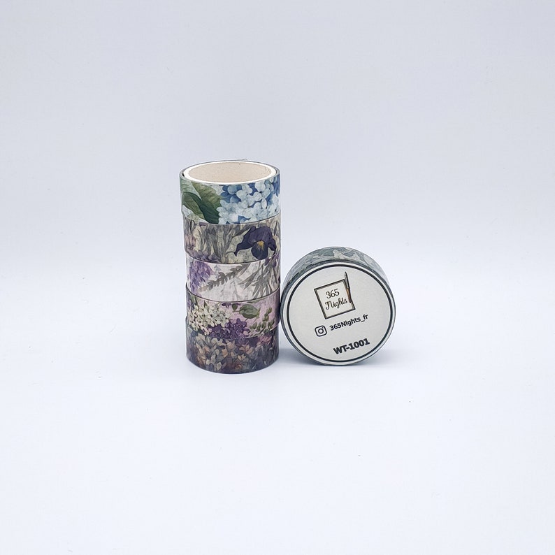 May include: A stack of four rolls of decorative washi tape featuring various floral and botanical patterns in shades of blue, purple, and green, alongside one roll showing a white label with the text 365 Nights, 365Nights_fr, and WT-1001.