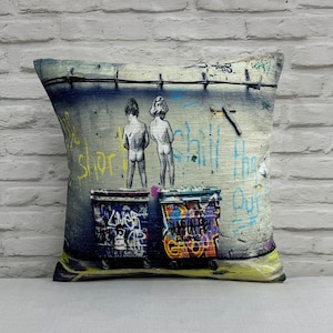 May include: A throw pillow with a graffiti-style design featuring two figures standing on top of garbage bins. The graffiti says "Chill Here Our" and "Shorty".