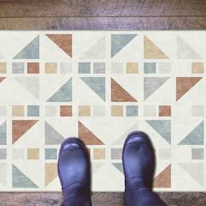 Puede incluir: Alfombra rectangular con un patrón geométrico de triángulos y cuadrados en tonos marrón, gris y azul sobre un fondo crema. La alfombra está sobre un suelo de madera, con dos pares de botas negras visibles.
