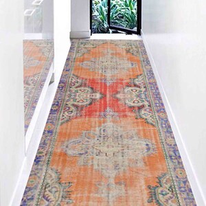 May include: A long, narrow, orange and red rug with a faded, vintage look. The rug has a detailed, intricate pattern with blue, green, and white accents. The rug is laid out in a hallway with white walls.
