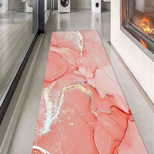 Op de afbeelding: Een roze en wit vloerkleed met gouden en witte abstracte marmerpatronen. Het kleed ligt op een lichtgrijze vloer.