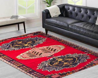 May include: A rectangular red rug with three central patterned designs in shades of blue, gold, and beige. The rug is placed on a light-colored wooden floor, with a black leather sofa and a bench in the background.