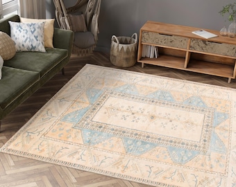 May include: A large, rectangular area rug with a cream-colored central field and a geometric border in shades of light blue and tan. The rug is placed on a wooden floor, and a green sofa and wooden cabinet are visible in the background.
