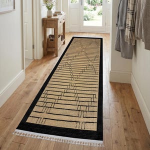Puede incluir: Una alfombra de pasillo rectangular larga con una textura tejida en color tostado natural y un patrón geométrico negro repetitivo. La alfombra está enmarcada por un borde negro sólido en todos sus lados y rematada con flecos blancos cortos.