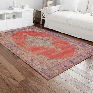 May include: A rectangular, distressed-look rug with a central medallion design. The rug features a vibrant red field with a faded, vintage appearance, bordered by a contrasting purple and beige pattern. The rug is placed on a hardwood floor.