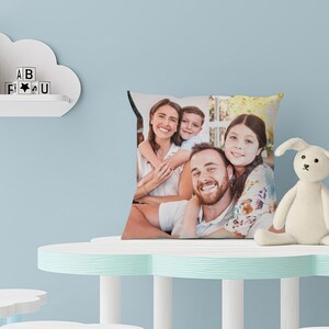 May include: Square pillow featuring a family portrait. The pillow displays a smiling family of four. A white stuffed bunny sits on a light blue table. A cloud-shaped shelf holds wooden shapes and letter blocks.