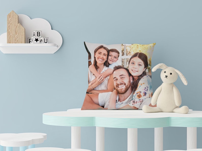 May include: Square pillow featuring a family portrait. The pillow displays a smiling family of four. A white stuffed bunny sits on a light blue table. A cloud-shaped shelf holds wooden shapes and letter blocks.
