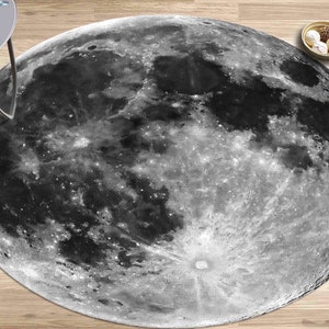 May include: A round rug featuring a detailed grayscale image of the moon's surface. The rug's design includes craters and textures, creating a realistic lunar landscape. The rug is placed on a wooden floor.