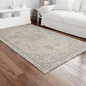 Puede incluir: Una alfombra beige claro con un patrón floral en tonos azules, rosas y crema. La alfombra tiene un aspecto desgastado y vintage, perfecta para añadir un toque de estilo bohemio a cualquier habitación.