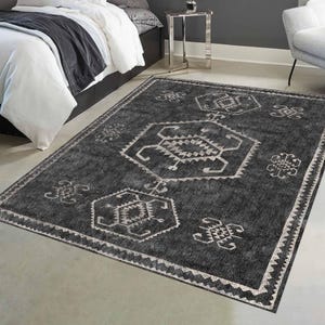 May include: A rectangular, dark gray area rug with a geometric pattern in white. The rug features a central design and repeating motifs along the border. The rug is placed on a concrete floor near a bed and a chair.