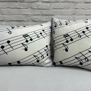 May include: Two white throw pillows with black musical notes printed on them. The pillows are on a white sofa in front of a white brick wall.