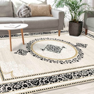 May include: A white area rug with a black and gold geometric pattern. The rug features a central medallion with a black and white floral design. The rug is on a wooden floor in a living room setting.